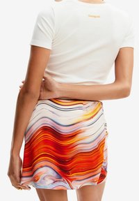 Vit ribbad kortärmad topp med orange logotyp, matchad med en livlig minikjol med vågmönster i röda, orange, lila och krämfärgade toner.