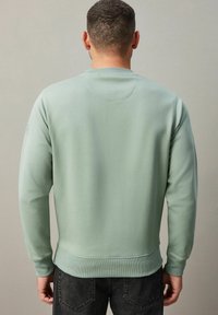 Sudadera verde claro con cuello redondo, puños y dobladillo acanalados. Textura suave con un logo en la manga izquierda. Llevada con jeans oscuros.