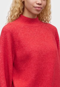Femme blonde portant un pull en laine rouge avec un col montant côtelé, vue des épaules au menton sur un fond blanc.