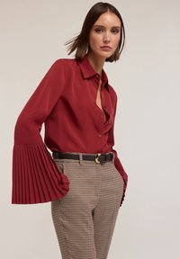 Blouse rouge à col avec des manches cloche plissées, associée à un pantalon à carreaux et une ceinture marron avec une boucle en or. Texture de tissu lisse.