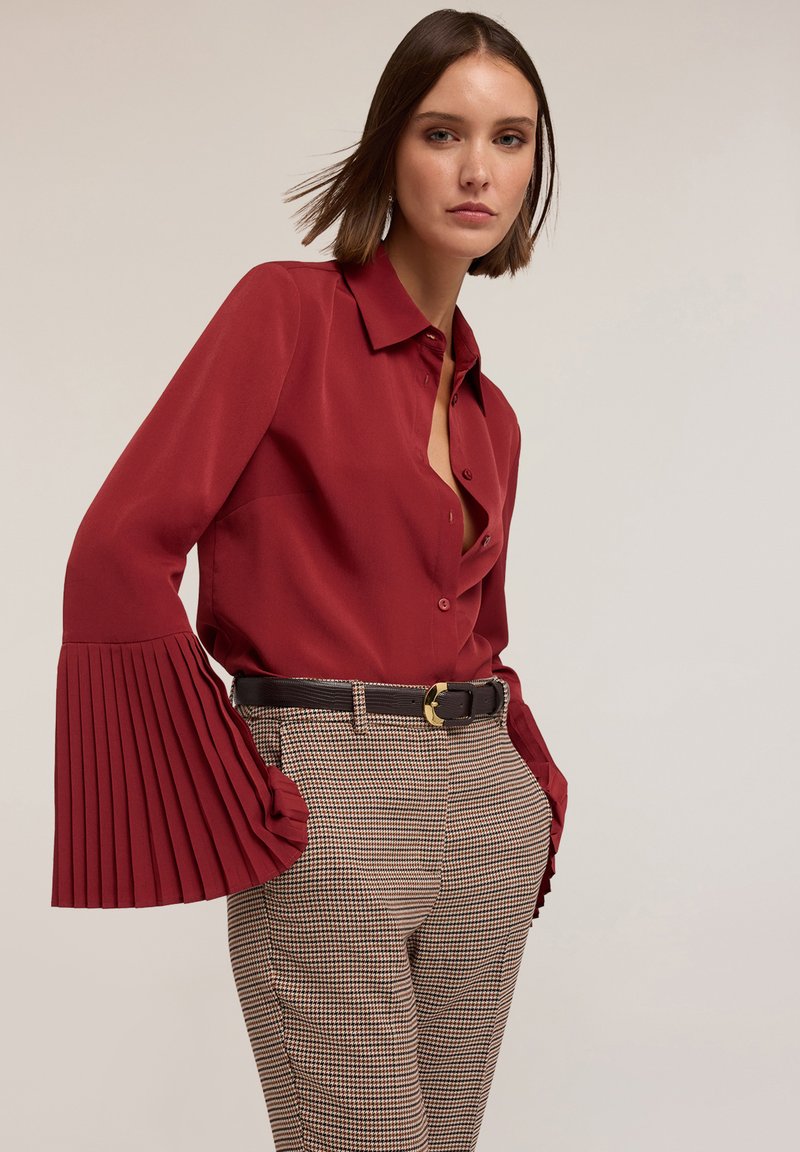 Blouse rouge à col avec des manches cloche plissées, associée à un pantalon à carreaux et une ceinture marron avec une boucle en or. Texture de tissu lisse.