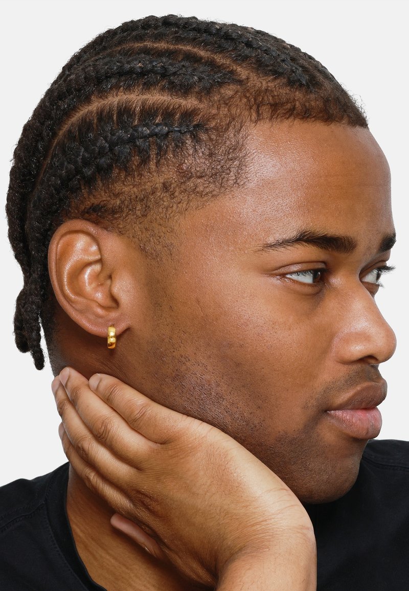 Boucle d'oreille en or sur un modèle avec des cheveux tressés. Forme légèrement courbée, texture lisse, attachée au lobe. Brillance subtile sur une chemise sombre.