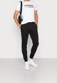 Vit t-shirt med "TOMMY JEANS" tryckt i svart och röd, tillsammans med svarta träningsbyxor och vita sneakers. Väskan är svart med ett rem.