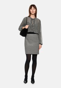 Schwarz-weiße gemusterte Bluse mit Rüschendetails, kombiniert mit einem taillierten schwarz-weiß karierten Rock. Model hält eine schwarze Handtasche.