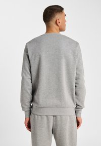 Mężczyzna z krótkimi włosami, ubrany w gładką, jasnoszaro szatę i pasujące szare dresy, stojący tyłem na tle białym.