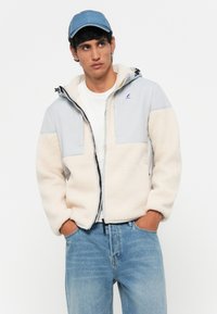Light beige fleece jacket with gray panels, zip closure, front pockets, and adjustable hood. Paired with a white t-shirt and blue jeans.