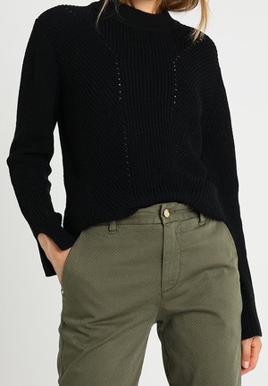 Schwarzer Strickpullover mit hohem Kragen, der strukturierte vertikale Streifen und offene Maschen-Details auf der Vorderseite aufweist. Kombiniert mit olivgrünen Hosen.