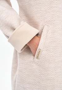 Hand in pocket of beige herringbone-patterned coat with ribbed cuff and beige "blue shadow" label on pocket edge.