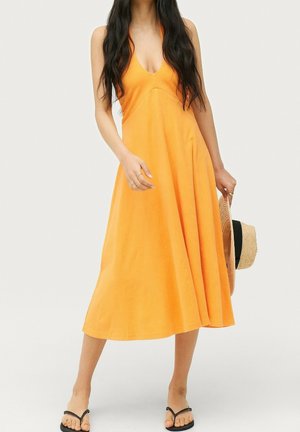 Kobieta w pomarańczowej, bezrękawnikowej sukience midi, trzymająca słomkowy kapelusz i czarne klapki, z długimi falowanymi włosami na jednolitym tle.