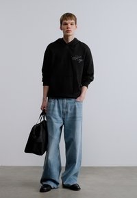 Μαύρο φούτερ με κουκούλα, μπροστινή τσέπη και διακριτικό λογότυπο, συνδυασμένο με ανοιχτό μπλε τζιν παντελόνι με φαρδιά μπατζάκια. Κρατάει μια μαύρη τσάντα. Λείο υλικό.