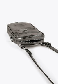 Malá čierna prešívaná kožená crossbody taška so strieborným zipsom, ktorý je čiastočne otvorený, a nastaviteľným popruhom na bielom pozadí.