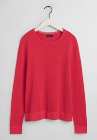 Pull tricoté rouge à manches longues avec texture et col rond, suspendu à un cintre blanc sur un fond gris uni.