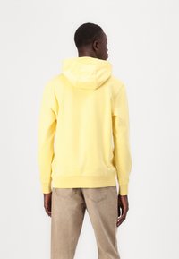 Gelber Kapuzenpullover mit weicher Textur, gerippten Bündchen und Saum. Getragen mit beigen strukturierten Hosen. Rückansicht vor einem schlichten weißen Hintergrund.