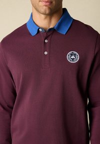 Bordowa koszula polo z długim rękawem, niebieskim kołnierzykiem, zapięciem na dwa guziki i okrągłym logo na lewej piersi. Wykonana z miękkiej mieszanki bawełny.