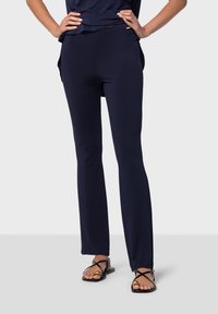 Mörkblå stretchbyxor med en figurnära siluett, sidofickor och en mjuk textur, kombinerat med svarta sandaler med guldaccent.