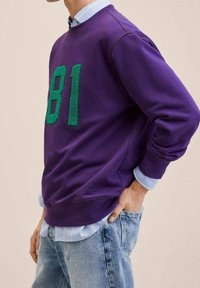 Sudadera morada con números texturizados verdes "81" en el frente. Presenta puños y dobladillo acanalados. Llevada sobre una camisa de cuello azul claro.
