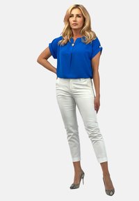 Niebieska bluzka z krótkim rękawem i detalem kołnierza, zestawiona z białymi dopasowanymi spodniami i wzorzystymi szpilkami. Gładki materiał, nowoczesny design.