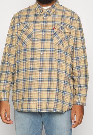 Uomo che indossa una camicia a quadri gialla, blu e rossa con due tasche sul petto con bottone e jeans azzurri.