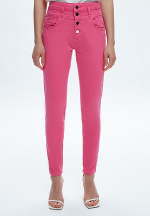Femme portant un pantalon rose taille haute, slim avec quatre boutons devant et des talons blancs à bouts ouverts, debout devant un fond uni.