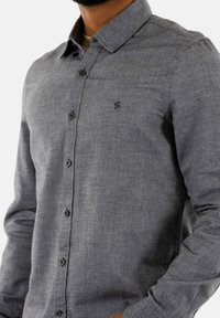 Camisa gris de manga larga y botones, hecha de una tela texturizada, con cuello, botones negros y un pequeño logo en el lado izquierdo del pecho.