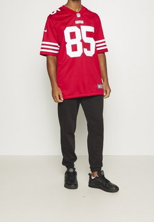 Maillot rouge de la NFL avec des rayures blanches et le numéro 85, associé à un pantalon de survêtement noir et des baskets noires. Le maillot présente un design en V.