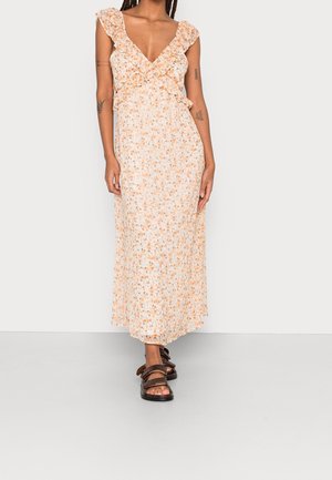 Vrouw draagt een mouwloze, V-hals, bloemen maxi-jurk met ruches, staat tegen een effen lichte achtergrond en draagt bruine sandalen.