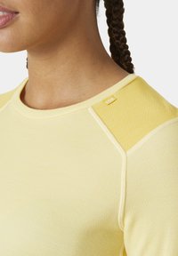Žlutý sportovní top s texturovanou tkaninou, kulatým výstřihem a kontrastním akcentem na rameni. Obsahuje malou nášivku s logem na límci.