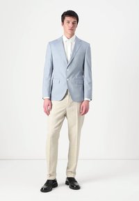 Svetlo modra blazer z dvema sprednjima gumboma, v kombinaciji s bež hlačami in črnimi čevlji. Gladek material in prilegajoč kroj poudarjata strukturo.