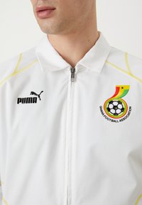 Hvid sportsjakke med gule detaljer, et sort Puma-logo og et emblem fra Ghanas fodboldforbund med et fodbolddesign.