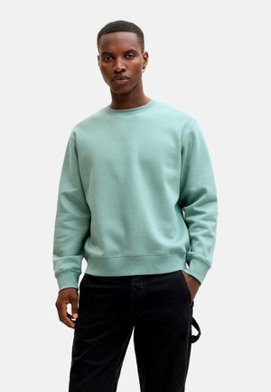 Mladi moški stoji, oblečen v svetlo zelen pulover in črne hlače, z eno roko v žepu, na enostavni ozadju.