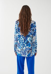 Blusa a maniche lunghe con motivo floreale blu, caratterizzata da tonalità di blu e bianco. Abbinata a pantaloni blu brillante, tessuto morbido, vestibilità rilassata.