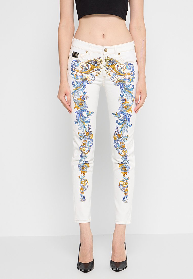 Weiße Slim-Fit-Jeans mit blauen und goldenen floralen Barockmustern, die vertikal an der Vorderseite verlaufen, getragen mit schwarzen Absatzschuhen und einem schwarzen Cropped-Top.