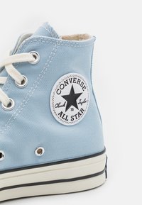 Sapatilha de cano alto em lona azul claro com atacadores brancos e um patch redondo com uma estrela preta, logotipo "Converse All Star" e sola de borracha.