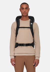 Black backpack with adjustable padded shoulder straps, a sternum strap, and a logo patch, worn over a light beige sweatshirt and brown pants.