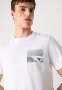 Hvid bomulds t-shirt med en frontlomme, der har et fotografisk print i gråtoner og "BOSS"-logoet nederst. Korte ærmer.