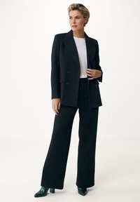 Blazer negro de doble botonadura sobre una camisa blanca, combinado con pantalones anchos negros y zapatos de tacón verde. Corte entallado, tejido suave.