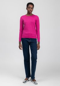 Kvinde iført en lys pink langærmet sweater, mørkeblå jeans og blanke sølv flade sko, stående mod en simpel lys grå baggrund.