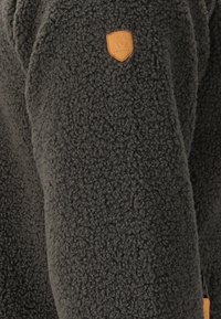 Fleece jacka i mörkgrått, med en plyschliknande textur, rund form och en läderlogotyp. Inkluderar dragkedja och sidoficka.