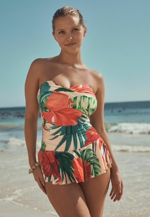 Kobieta stoi na plaży w suknie kąpielowej bez ramiączek z czerwonymi i zielonymi tropikalnymi wzorami, delikatnie uśmiechając się do kamery.