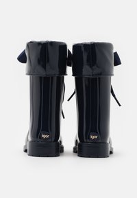 Bottes de pluie en caoutchouc brillantes bleu marine avec un bout rond, nœuds en biais en haut et une semelle texturée, arborant un logo "igor" doré sur le talon.