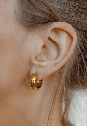 Goldene Creolen mit einer glänzenden, glatten Oberfläche, die in einer Nahaufnahme eines Ohrs präsentiert werden und ihre kreisförmige Form sowie das polierte Finish hervorheben.