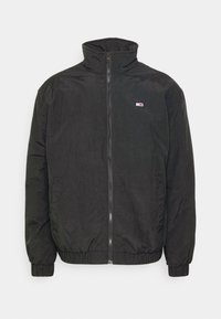 Giacca a vento nera con colletto alto e zip frontale. Presenta polsini e orlo elastici, e una piccola toppa con logo sul petto. Texture morbida.