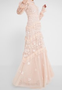 Lång, blek persikofärgad klänning med lager av tyll, blommig brodyr och paljetter. Har volangärmar och hög hals.