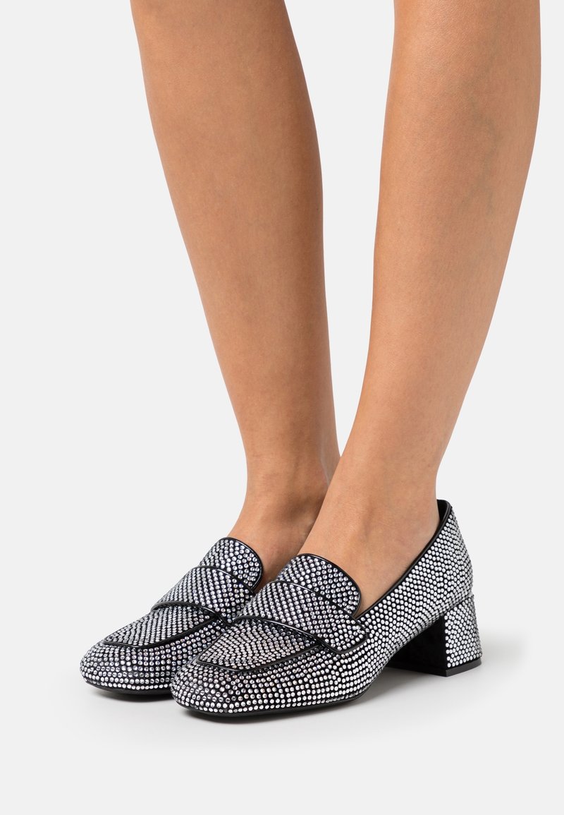 Jambes féminines portant des mocassins noirs à talon bloc recouverts de strass argentés sur un fond blanc.