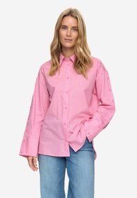 Roze gestreept overhemd met knopen, lange mouwen en grote manchetten. Voorzien van subtiele contrasterende strepen en een ontspannen pasvorm. Gecombineerd met lichtblauwe spijkerbroek.