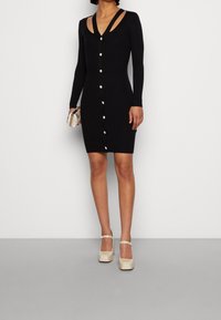 Abito in maglia nera a coste con maniche lunghe, scollatura a V e chiusura frontale con bottoni; presenta un design con spalle scoperte. Accompagnato da una pochette.