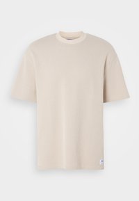 Beige tričko s krátkým rukávem, žebrované, s kulatým výstřihem a malou bílou etiketou u lemu na bílém pozadí.