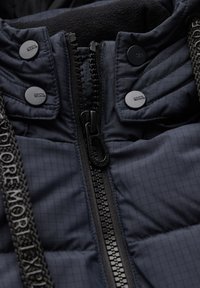 Chaqueta acolchada en color azul marino que presenta un cierre central con cremallera, botones a presión negros y una tela texturizada con un acento de marca en las correas.