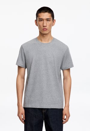 Jeune homme aux cheveux courts portant un t-shirt gris uni à manches courtes et un pantalon foncé, debout devant un fond blanc.