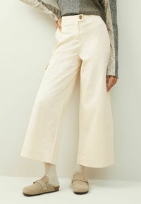 Wijduitlopende, crèmekleurige broek van lichtgewicht stof, met een hoge taille, sluiting met een knoop aan de voorkant en zijzakken, gecombineerd met beige klompen.